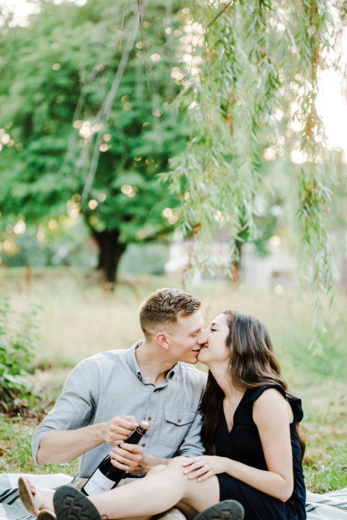 couple kissing during engagement session he is opening bottle of champagne 