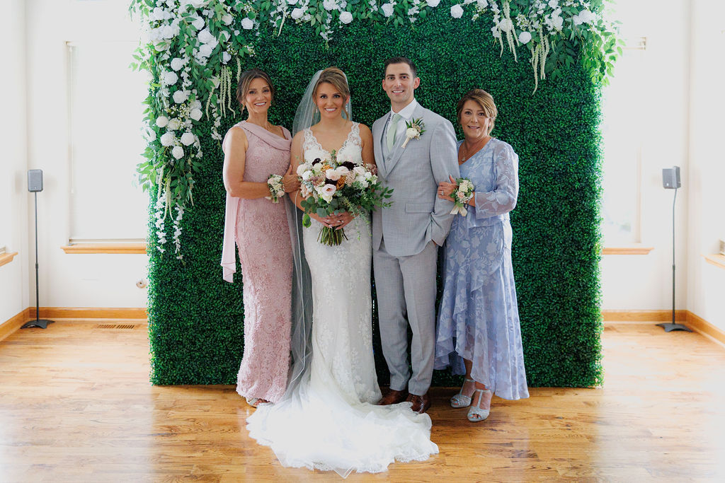 Bride and groom standing between their two mothers, all smiling warmly at the camera.